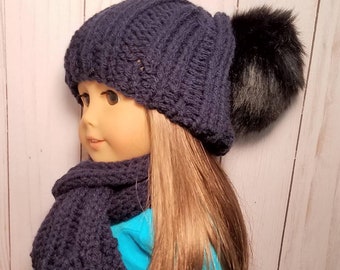 Puede incluir: Un gorro de punto azul marino con un pompón de piel sintética negra y una bufanda a juego. El gorro y la bufanda están hechos para una muñeca.