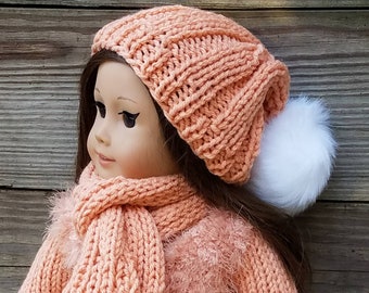 Puede incluir: Un conjunto de gorro y bufanda de punto de color melocotón con un pompón blanco en el gorro. El conjunto está hecho para una muñeca.