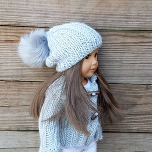 Puede incluir: Un gorro de punto azul claro con un pomp&oacute;n esponjoso en la parte superior. El gorro lo lleva una mu&ntilde;eca con el pelo largo y casta&ntilde;o y un c&aacute;rdigan de punto azul claro.