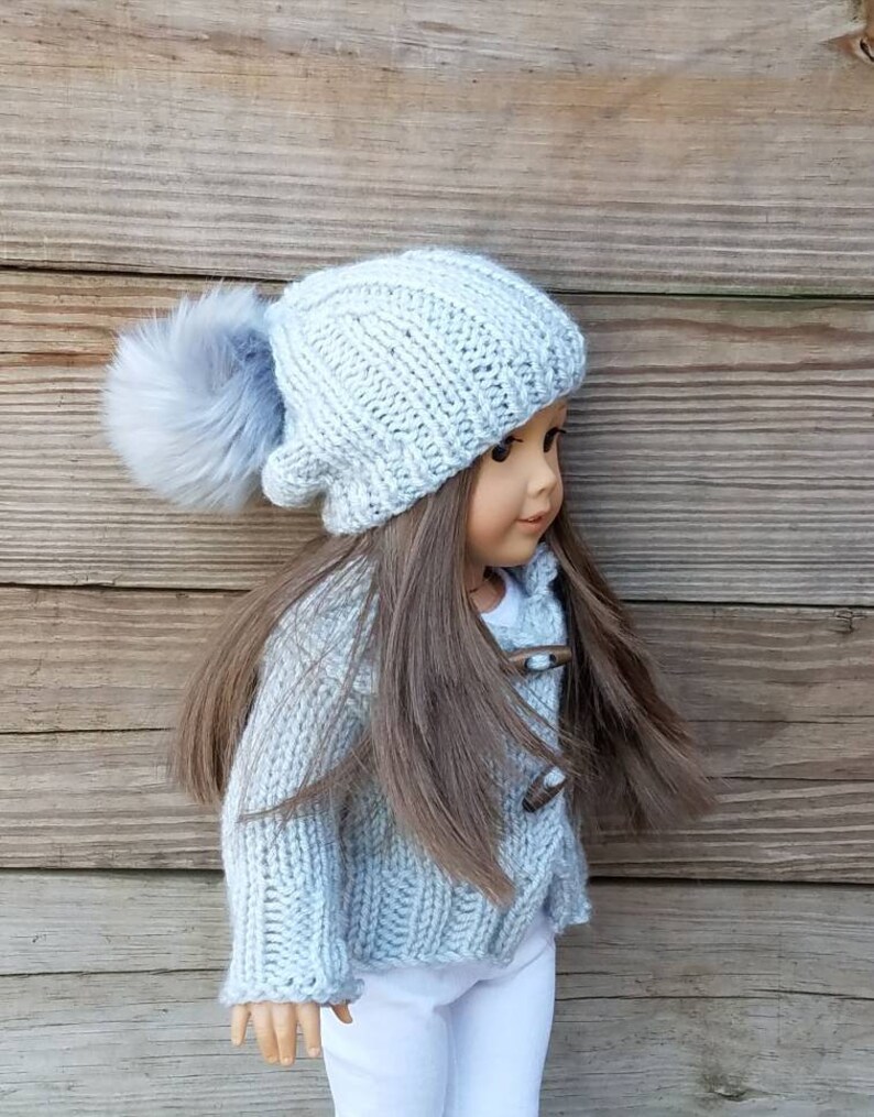 Puede incluir: Un gorro de punto azul claro con un pomp&oacute;n esponjoso en la parte superior. El gorro lo lleva una mu&ntilde;eca con el pelo largo y casta&ntilde;o y un c&aacute;rdigan de punto azul claro.
