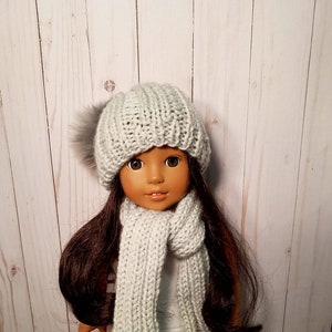 Puede incluir: Una mu&ntilde;eca con un gorro de punto gris con un pomp&oacute;n esponjoso y una bufanda a juego en gris.