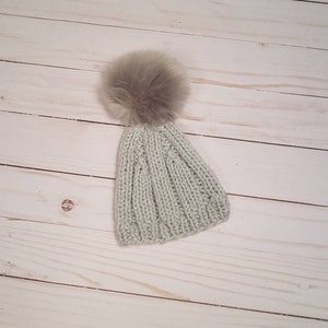 Puede incluir: Un gorro de punto gris claro con un pomp&oacute;n marr&oacute;n esponjoso en la parte superior.