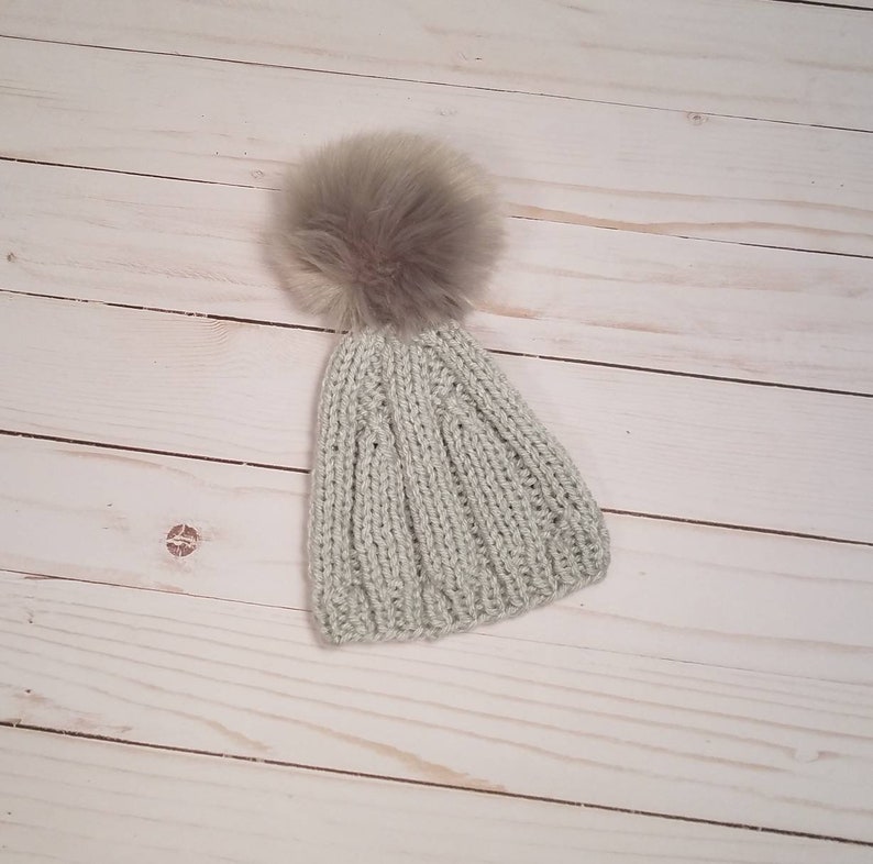 Puede incluir: Un gorro de punto gris claro con un pomp&oacute;n marr&oacute;n esponjoso en la parte superior.
