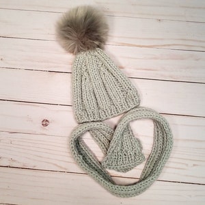 Puede incluir: Un gorro de punto verde claro con un pomp&oacute;n marr&oacute;n esponjoso y una bufanda a juego.