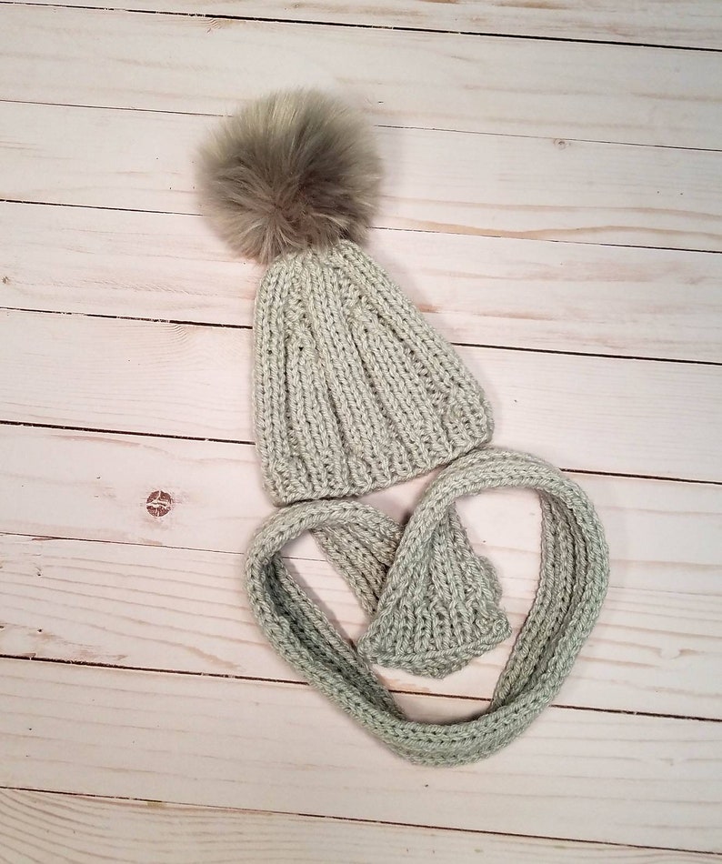 Puede incluir: Un gorro de punto verde claro con un pomp&oacute;n marr&oacute;n esponjoso y una bufanda a juego.