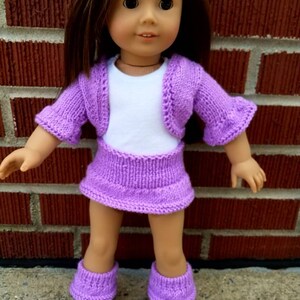 May include: A purple knitted doll outfit with a cardigan, skirt, and boots. The doll is wearing a white shirt underneath the cardigan.