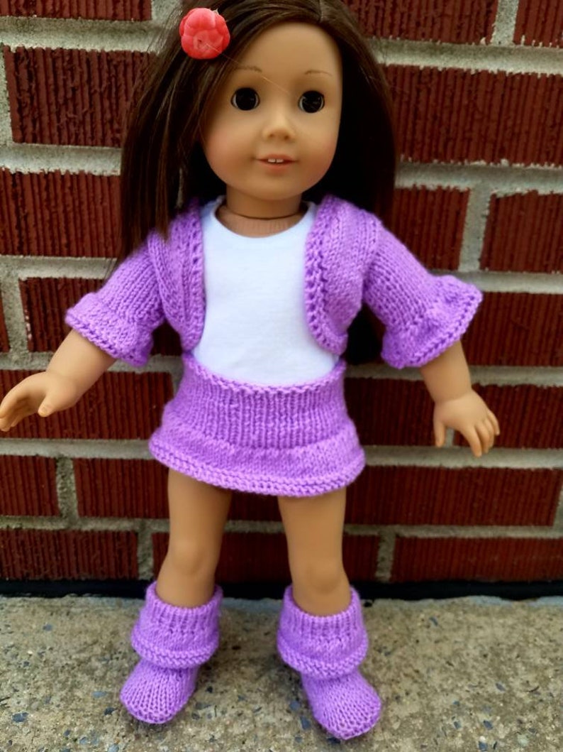 May include: A purple knitted doll outfit with a cardigan, skirt, and boots. The doll is wearing a white shirt underneath the cardigan.