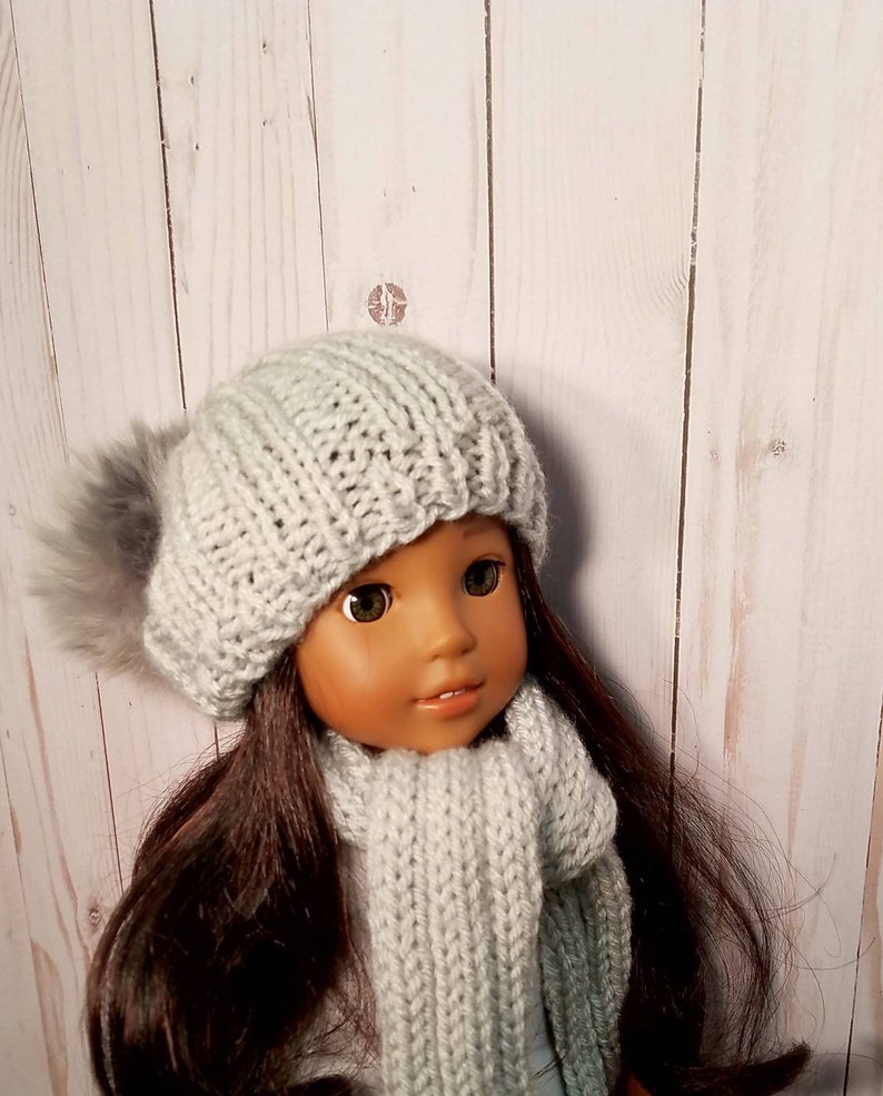 Puede incluir: Una mu&ntilde;eca con un gorro de punto gris con un pomp&oacute;n esponjoso y una bufanda a juego de color gris.