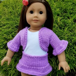 May include: A purple knitted doll outfit with a cardigan and skirt. The doll is wearing a white undershirt and has brown hair with a pink flower.
