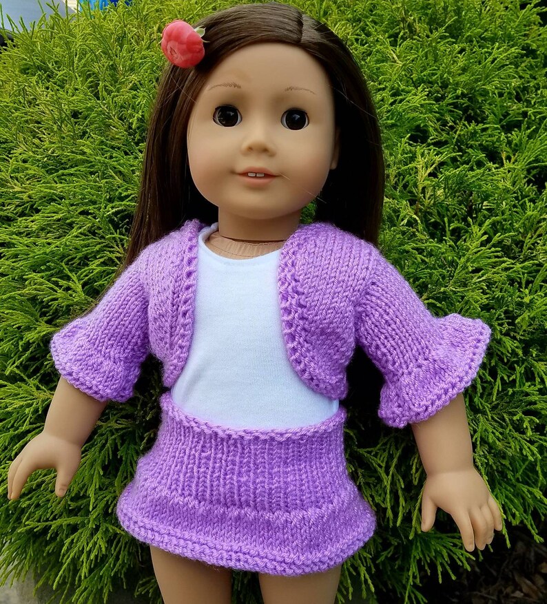 May include: A purple knitted doll outfit with a cardigan and skirt. The doll is wearing a white undershirt and has brown hair with a pink flower.