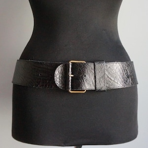 May include: A wide black belt with a textured, embossed pattern and a gold-colored rectangular buckle. The belt is displayed on a black mannequin, showcasing its design and fit. The belt is a fashion accessory.