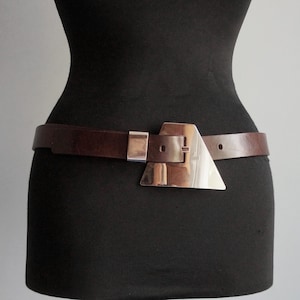 Op de afbeelding: Een bruine leren riem met een zilverkleurige, geometrische gesp. De riem wordt gedragen rond een zwarte mannequin, die het ontwerp en de pasvorm laat zien. De gesp heeft een driehoekige vorm en een rechthoekige sluiting.