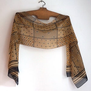 May include: A patterned scarf in shades of brown and black, displayed on a wooden hanger. The scarf has a geometric design with stripes, ovals, and dots. The ends of the scarf are a darker black colour. The scarf is made of a sheer material.