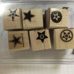 Puede incluir: Un juego de seis sellos de madera con diferentes diseños de estrellas. Los sellos están en un contenedor de plástico transparente. Los sellos son en blanco y negro y tienen un aviso de copyright en la parte inferior.