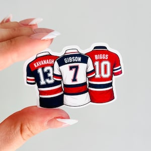 May include: Three sticker decals of hockey jerseys. The first jersey is navy blue, red, and white with the name "Kavanagh" and the number "13". The second jersey is white, navy blue, and red with the name "Gibson" and the number "7". The third jersey is red, white, and navy blue with the name "Biggs" and the number "10".