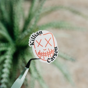 May include: White sticker with a red outline of a smiling face with X's for eyes. The text "Killian Carson" is written around the face.
