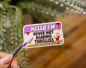 Könnte beinhalten: Ein lila-weißer Aufkleber mit dem Text "Hello I'm Never Not Reading Thrillers" mit einem blutigen Handabdruck und einem blutigen Messer. Der Aufkleber befindet sich auf einer Holzoberfläche.
