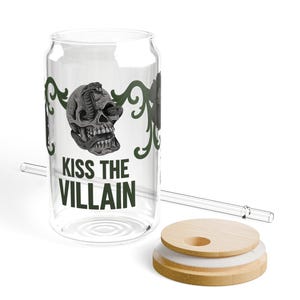 May include: Clear glass can-shaped drinking glass with a skull and snake design, the text "KISS THE VILLAIN", a wooden lid, and a clear straw. The skull is gray and the text is black. Green decorative flourishes are on the glass.