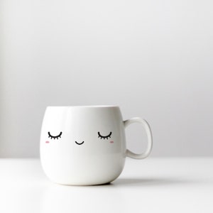 K&ouml;nnte beinhalten: Wei&szlig;e Keramiktasse mit einem niedlichen Comic-Gesicht mit geschlossenen Augen und einem L&auml;cheln.