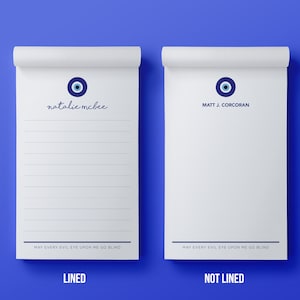 May include: Two white notepad designs with a blue evil eye symbol and the text "MAY EVERY EVIL EYE UPON ME GO BLIND". One notepad is lined and the other is not lined. The text "LINED" and "NOT LINED" is printed below each notepad.