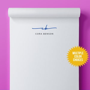 May include: A white notepad with a blue handwritten signature that says "cb" and the text "CORA BENSON" below the signature. A yellow starburst sticker with the text "MULTIPLE COLOR CHOICES" is on the right side of the notepad.