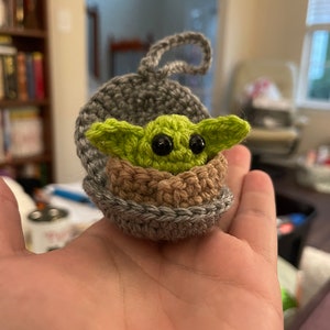 May include: A small crocheted green and brown Yoda character in a gray crocheted pod with a loop for hanging.