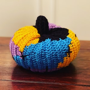 May include: A crocheted pumpkin with a black stem and a patchwork design of yellow, blue, purple, and black yarn.