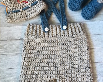 Puede incluir: Un conjunto de ropa de bebé tejido a crochet en color beige y azul. El conjunto incluye un gorro, un mono y botines. El mono tiene un diseño de tirantes cruzados y botones blancos. Los botines son de color azul y beige. El conjunto está hecho con un hilo jaspeado.