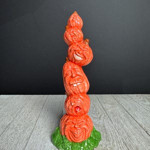 Könnte beinhalten: Gestapelte orangefarbene Keramik-Kürbis-Figur mit geschnitzten Gesichtern, auf einem grünen Sockel. Die Halloween-Dekoration hat ein skurriles Design mit einem Stapel Kürbisse, jeder mit einem einzigartigen Ausdruck.