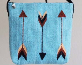 May include: A light blue woven crossbody bag featuring a pattern of three vertical arrows in shades of brown, black, and tan. The bag has a black leather strap knotted at the top and a silver-toned zipper pull shaped like a human hand.