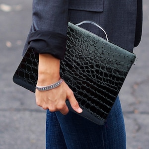 May include: A black crocodile-embossed clutch with a silver bracelet on the wrist of the person holding it.