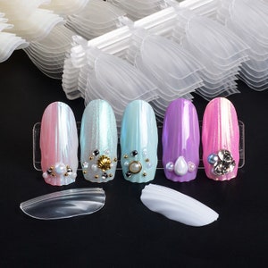 Op de afbeelding: Vijf gedecoreerde kunstnagels met iriserende afwerkingen in roze, blauw, paars en wit. De nagels zijn versierd met parels, schelpen en strass steentjes. De nagels worden gepresenteerd op een transparante kunststof standaard. Er zijn ook twee transparante kunststof nageltips in de afbeelding.