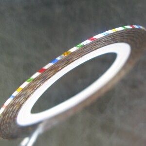 May include: A roll of silver nail art tape with multi-colored stripes. The tape is on a white backing.
