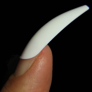 May include: A white, long, curved, artificial nail tip is attached to a finger.