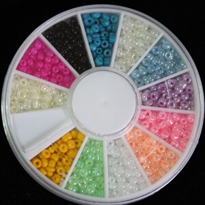 Pode incluir: Uma roda de pl&aacute;stico transparente com 12 compartimentos, cada um cheio de uma cor diferente de pequenas contas redondas. As contas s&atilde;o em tons de branco, rosa, azul, verde, amarelo e preto.