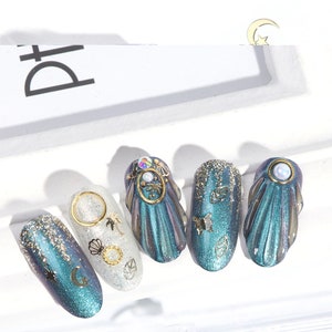 Op de afbeelding: Vijf tealblauwe en zilveren, iriserende nageltips met gouden glitter en verschillende gouden en zilveren versieringen, waaronder een halve maan, een ster, een schelp, een palmboom, een vlinder en bladeren.