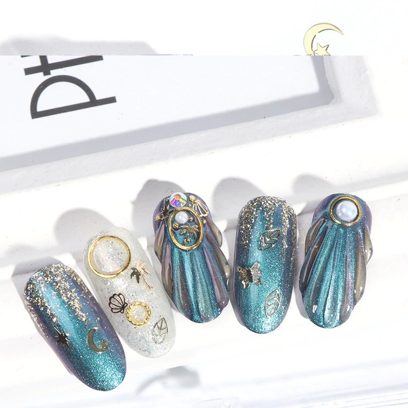 Op de afbeelding: Vijf tealblauwe en zilveren, iriserende nageltips met gouden glitter en verschillende gouden en zilveren versieringen, waaronder een halve maan, een ster, een schelp, een palmboom, een vlinder en bladeren.