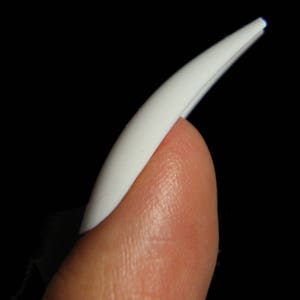 May include: A white plastic nail tip, curved and pointed, is held on a finger.