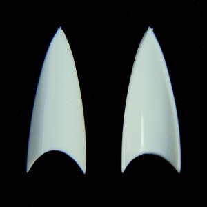 Puede incluir: Dos puntas de u&ntilde;as postizas en forma de estilete, de color blanco roto, se muestran sobre un fondo negro. Las puntas son puntiagudas y tienen un acabado liso y brillante. La base de cada u&ntilde;a tiene forma curva.