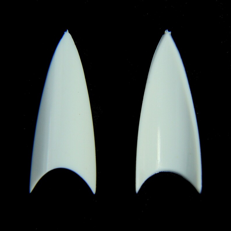 Puede incluir: Dos puntas de u&ntilde;as postizas en forma de estilete, de color blanco roto, se muestran sobre un fondo negro. Las puntas son puntiagudas y tienen un acabado liso y brillante. La base de cada u&ntilde;a tiene forma curva.