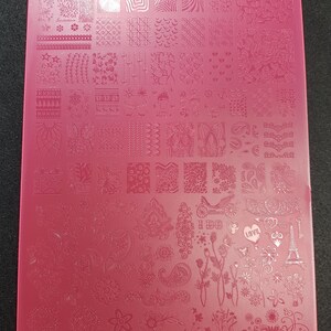 May include: A pink rectangular nail art stamping plate with etched designs. The plate features patterns including flowers, hearts, paisley, and geometric shapes. The plate is made of a smooth, glossy material for creating nail art.