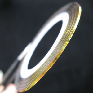May include: Gold nail art tape with a white backing. The tape is rolled up and the gold side is facing the camera.