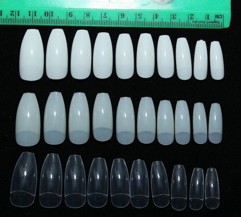 May include: Three rows of white and clear plastic false nails in various sizes. The nails are arranged in rows from smallest to largest. A ruler is visible at the top of the image.