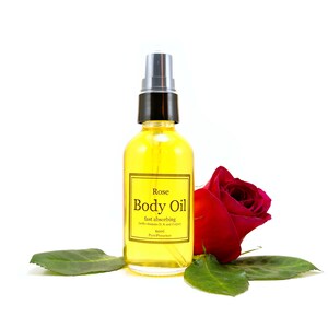 May include: A clear glass bottle of Rose Body Oil with a black spray top. The bottle is labeled with the text "Rose Body Oil fast absorbing (with vitamins D, E and C)" and "60ml PurePresence". The bottle is sitting on a white surface with a red rose and green leaves.