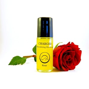 Könnte beinhalten: Eine gelbe Roll-on-Flasche mit Charged Mineralisierendes Deodorant im Duft Rose. Die Flasche hat einen schwarzen Deckel und ein weißes Wellenlogo. Im Hintergrund befindet sich eine rote Rose.