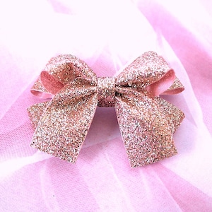 Könnte beinhalten: Eine roségoldene Glitzer-Schleife, mittig im Bild, vor einem zarten rosa Hintergrund. Die Schleife besteht aus einem glitzernden Material, mit einer klassischen Schleifenform. Der Hintergrund ist hellrosa, möglicherweise Tüll oder ein ähnlicher Stoff.
