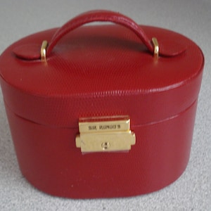 May include: An oval-shaped red jewelry case with a textured surface and a top handle. The case features a gold-toned metal latch on the front with the text SIR RONDO'S engraved on the clasp. The lid is secured with gold-toned metal hardware.