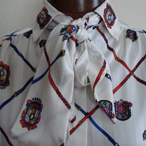 May include: White blouse with a repeating pattern of red and blue belts and crests. The blouse has a necktie and long sleeves. The pattern is set against a white background. The blouse is made of a silky material.