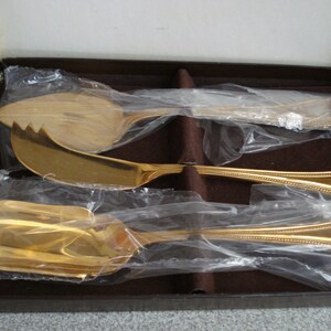 May include: Three gold-colored serving utensils in a brown box. The utensils are a butter knife, a cheese knife, and a serving spoon. The utensils are wrapped in plastic.