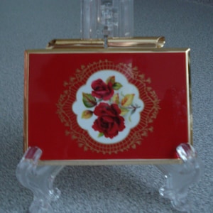 May include: A red compact with a gold trim and a white floral design featuring red roses.